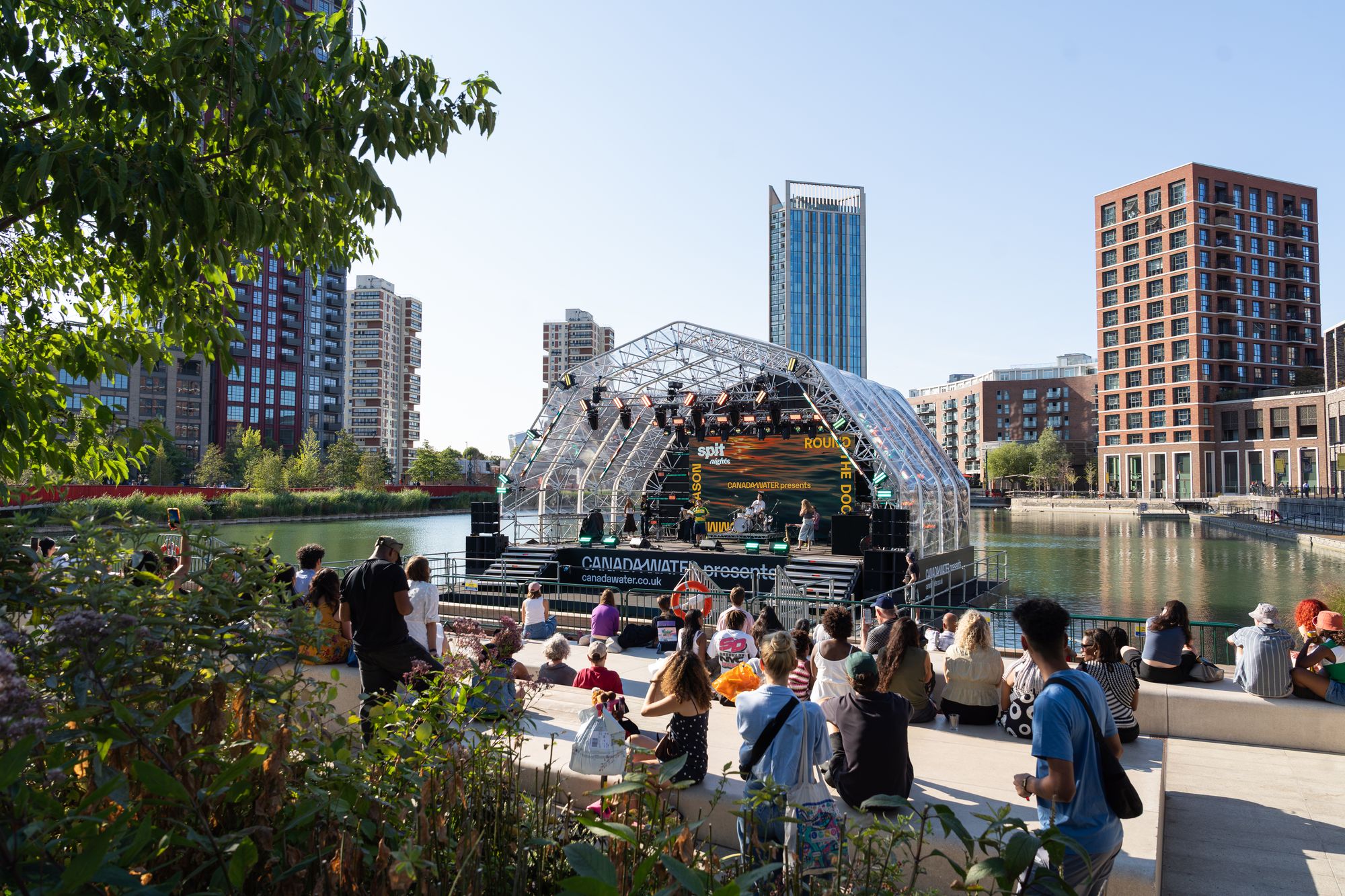 The floating stage on Canada Dock seen across the water, with the audience on the planted dockside.