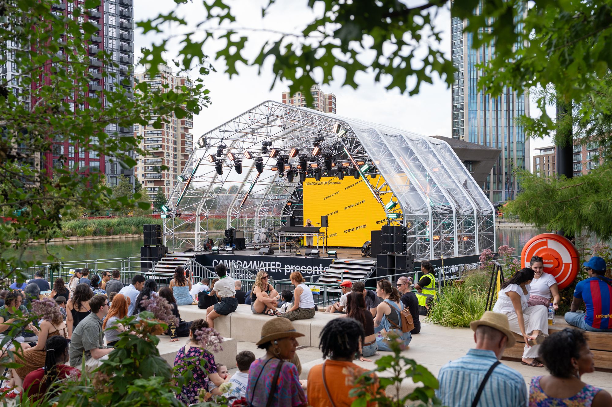 The floating stage and dockside crowd seen through the trees at Canada Water — the nature-inclusive setting of Round the Dock.