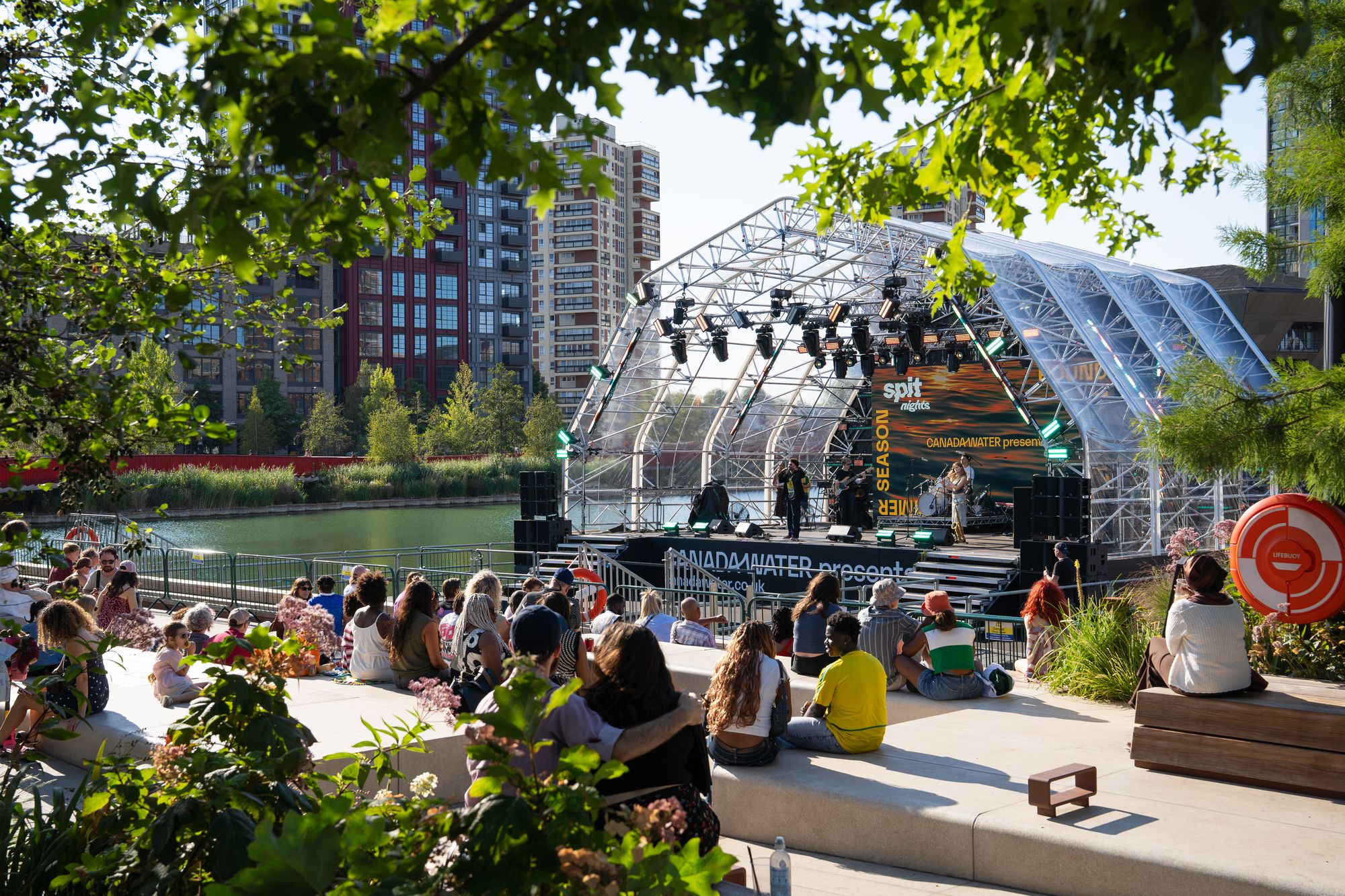 The Spit Night audience on the dockside steps, with the floating stage seen through the trees at Canada Water, London SE16.