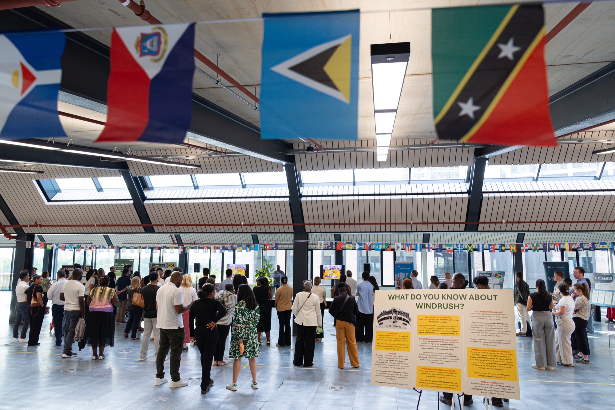 Caribbean national flags above guests and a Windrush information board at the celebration in the Dock Shed, Canada Water.