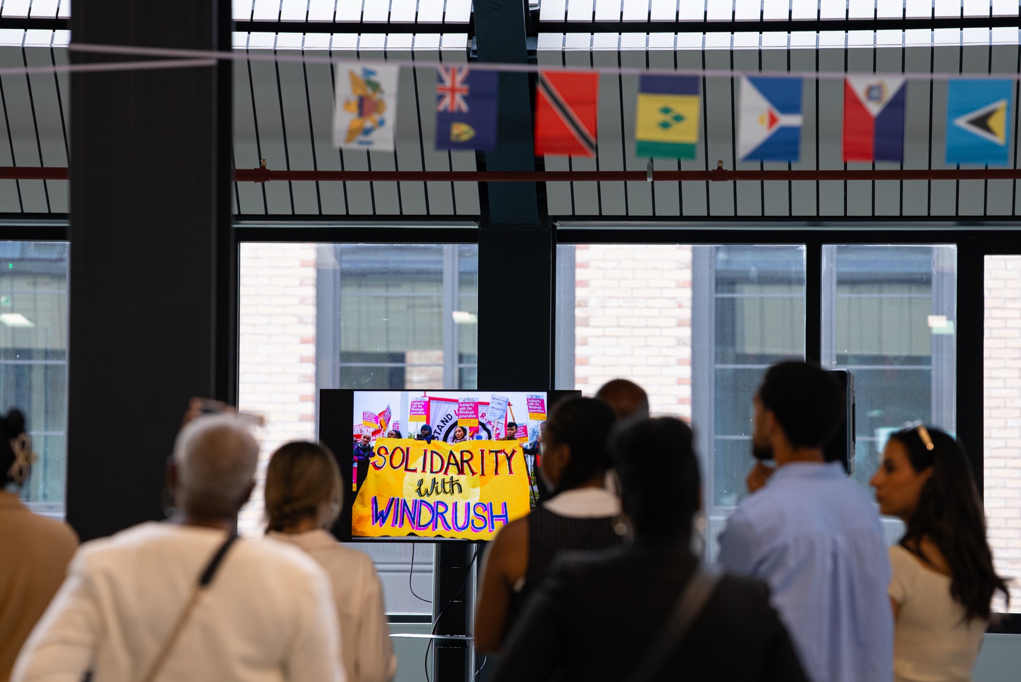 A "Solidarity with Windrush" screen beneath Caribbean flags as guests gather at the Dock Shed, Canada Water.