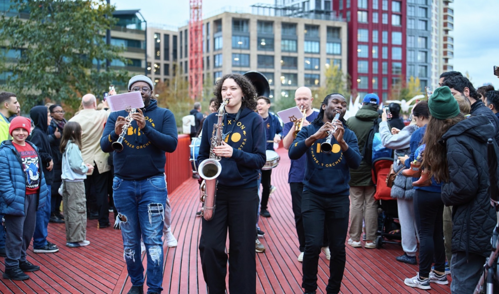 The Brixton Chamber Orchestra leads the opening procession across the boardwalk, drawing the crowd along the vivid red structure