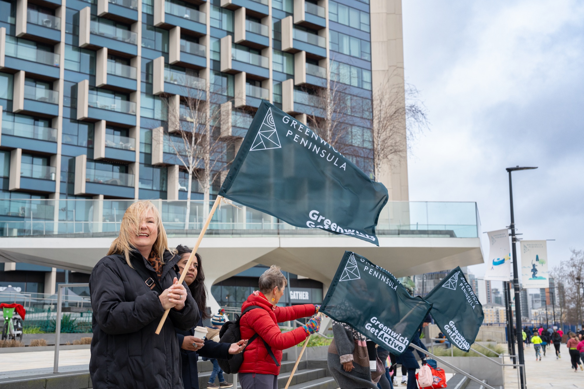 Volunteer marshals waving Greenwich Peninsula flags — the parkrun's community energy against the neighbourhood's distinctive residential architecture.