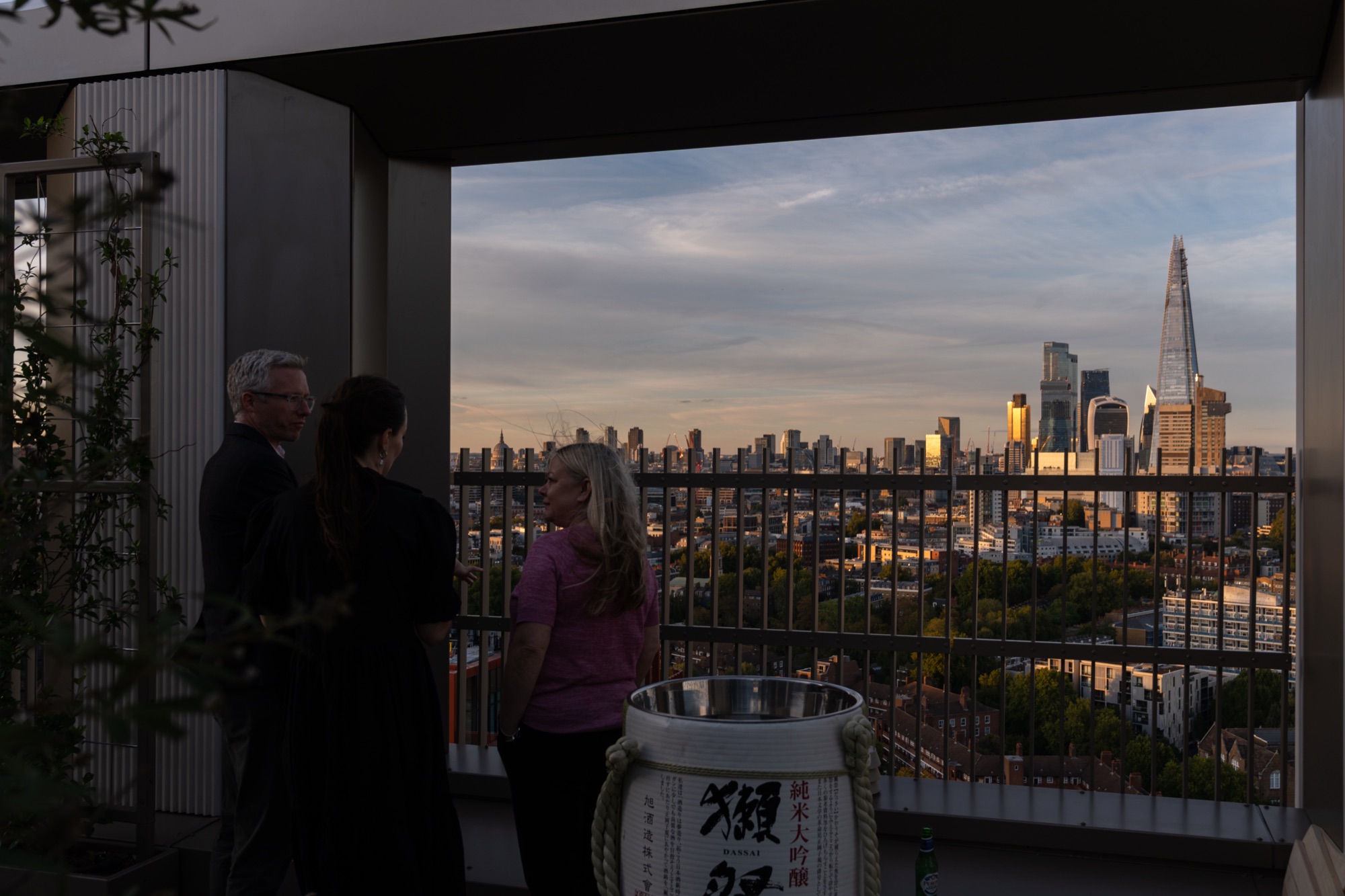 Guests on the Arbour Heights terrace at dusk, the sake barrel beside them and the London skyline beyond.