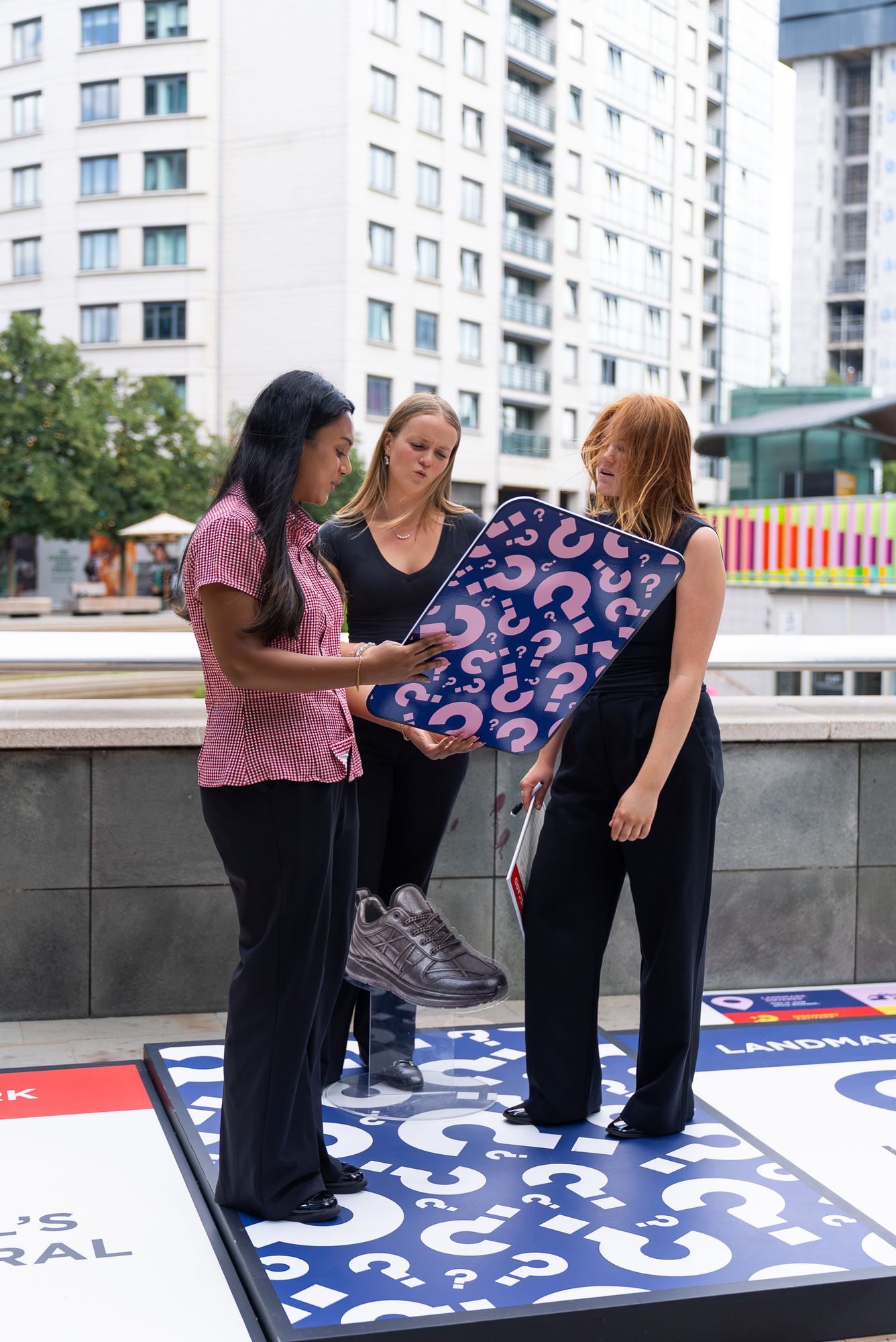 Three players reading a challenge card on a question-mark square of the giant board, Paddington Central.